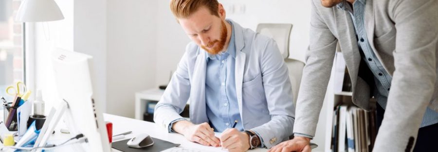 Two professionals, one writing on his note pad and one looking at the computer discussing about business transformation services